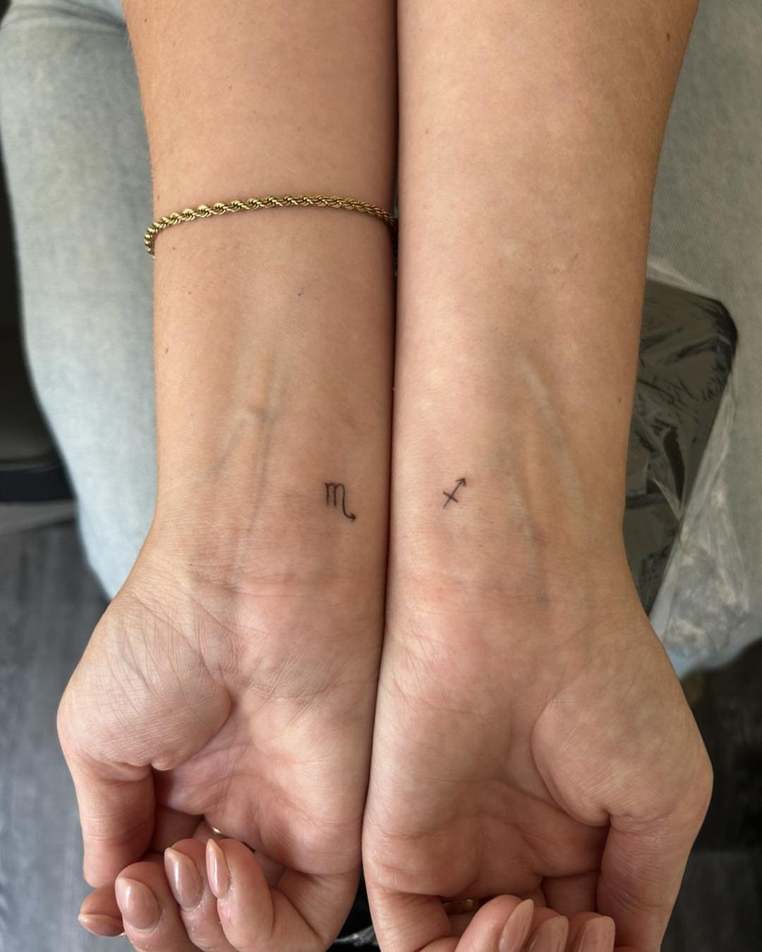 Two wrists with small black fine line tattoos of a Scorpio symbol and arrow.