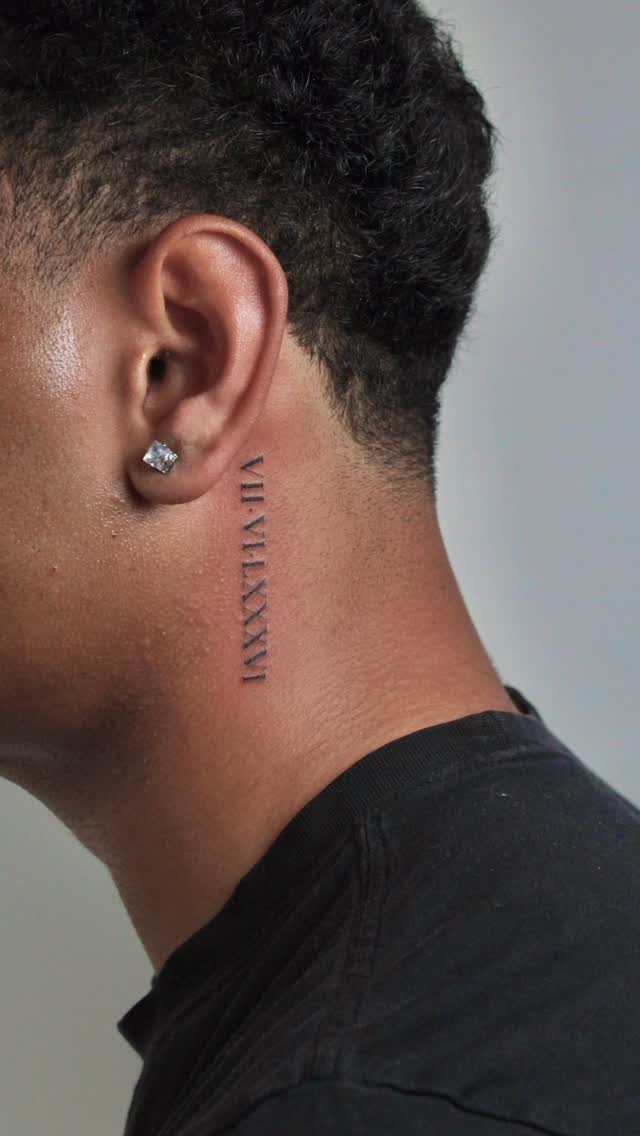 Man's neck featuring a vertical fine line Roman numeral date tattoo behind the ear.