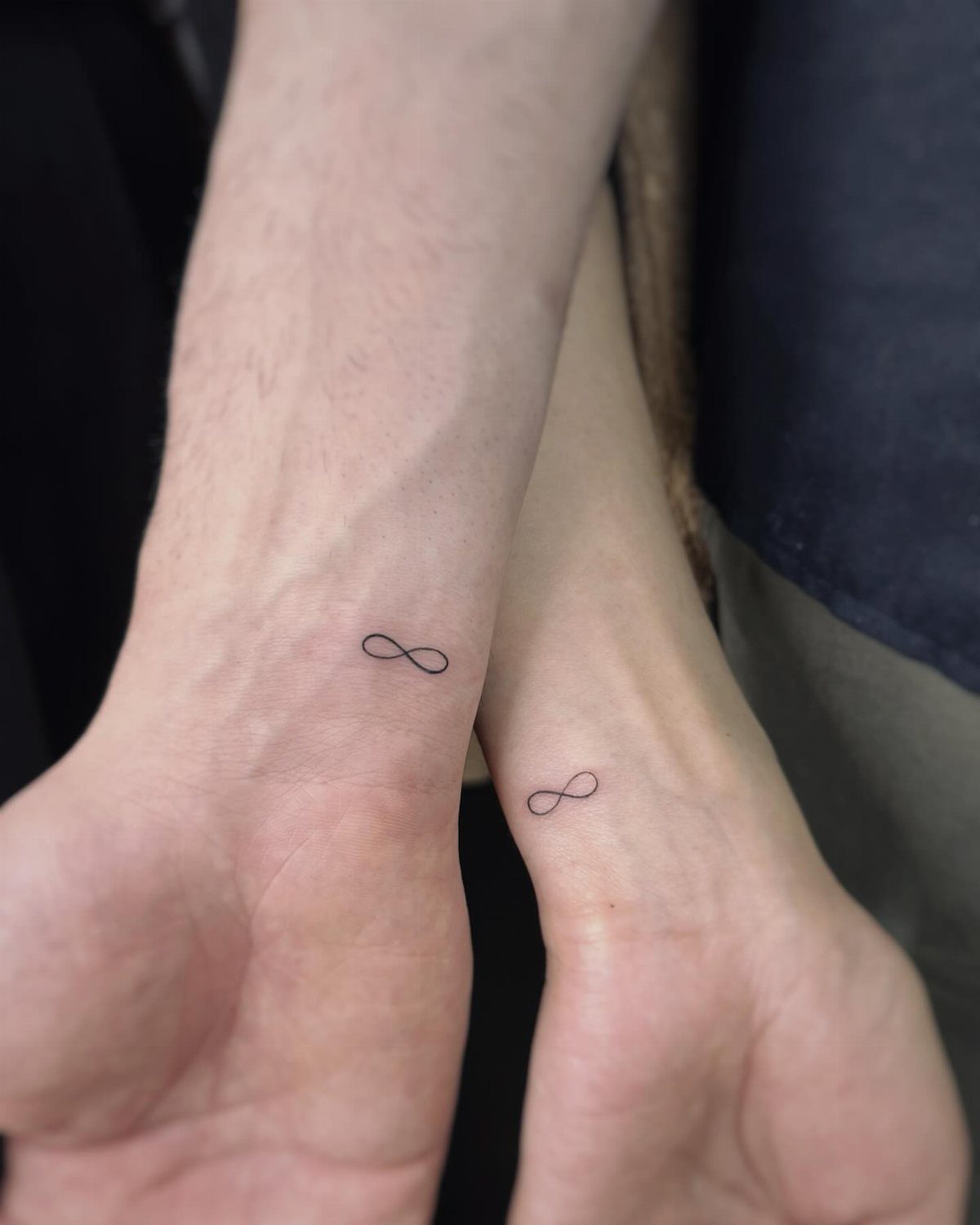 Two inner wrists featuring small, black fine-line infinity symbol tattoos side-by-side.