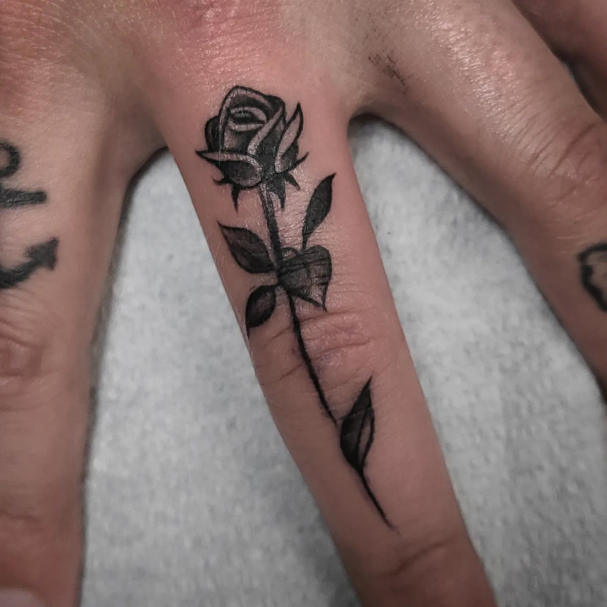 Black ink rose tattoo with stem and leaves on a person's middle finger.
