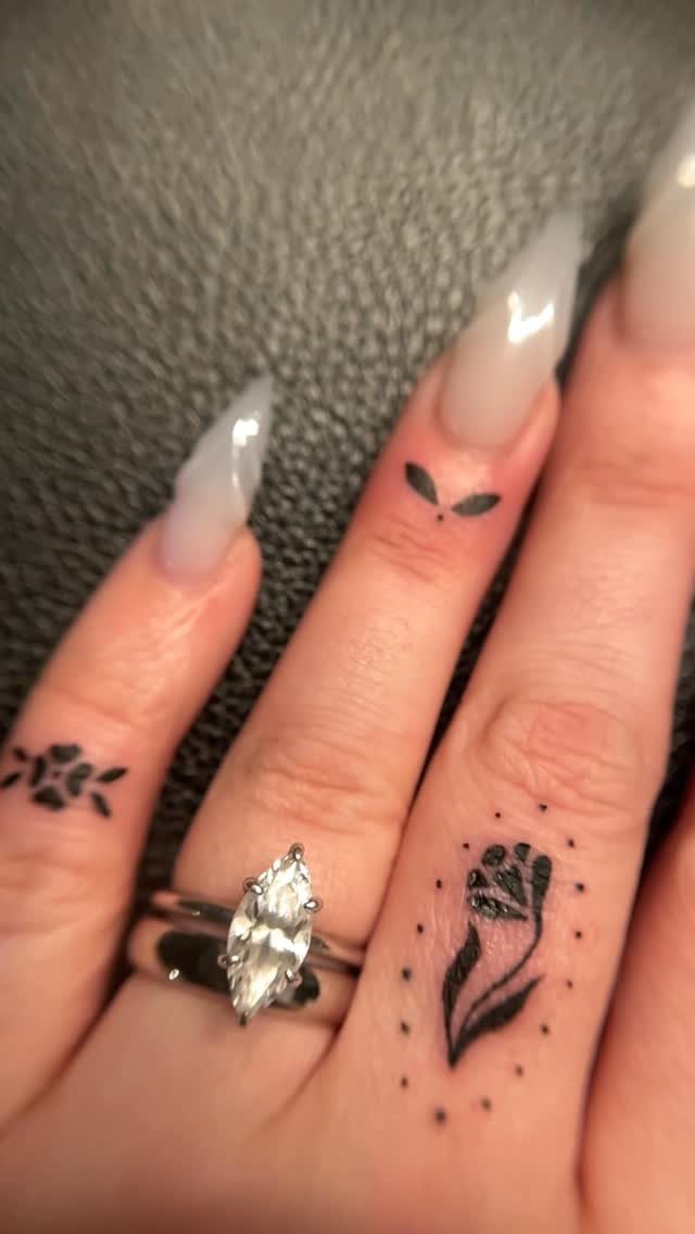 Small black rose and leaf tattoos on a person's fingers with rings.