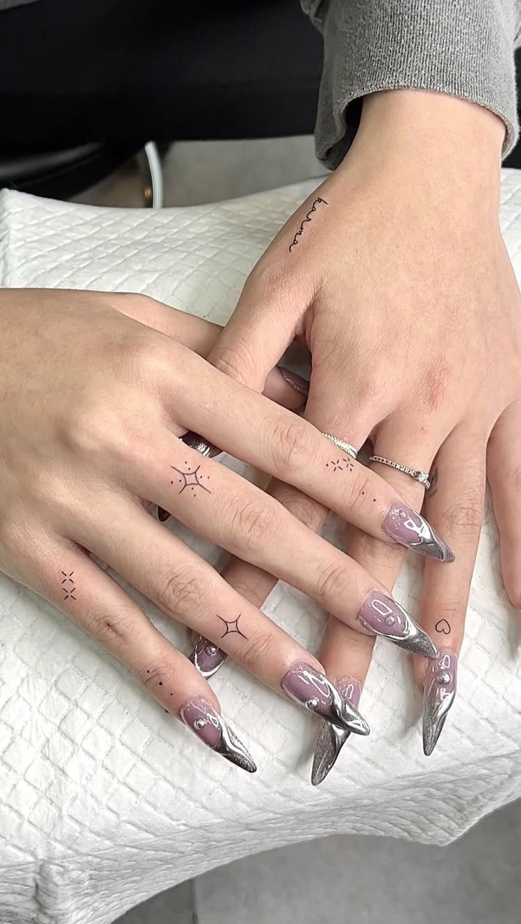 Hands with long metallic nails and small black star tattoos on the fingers.