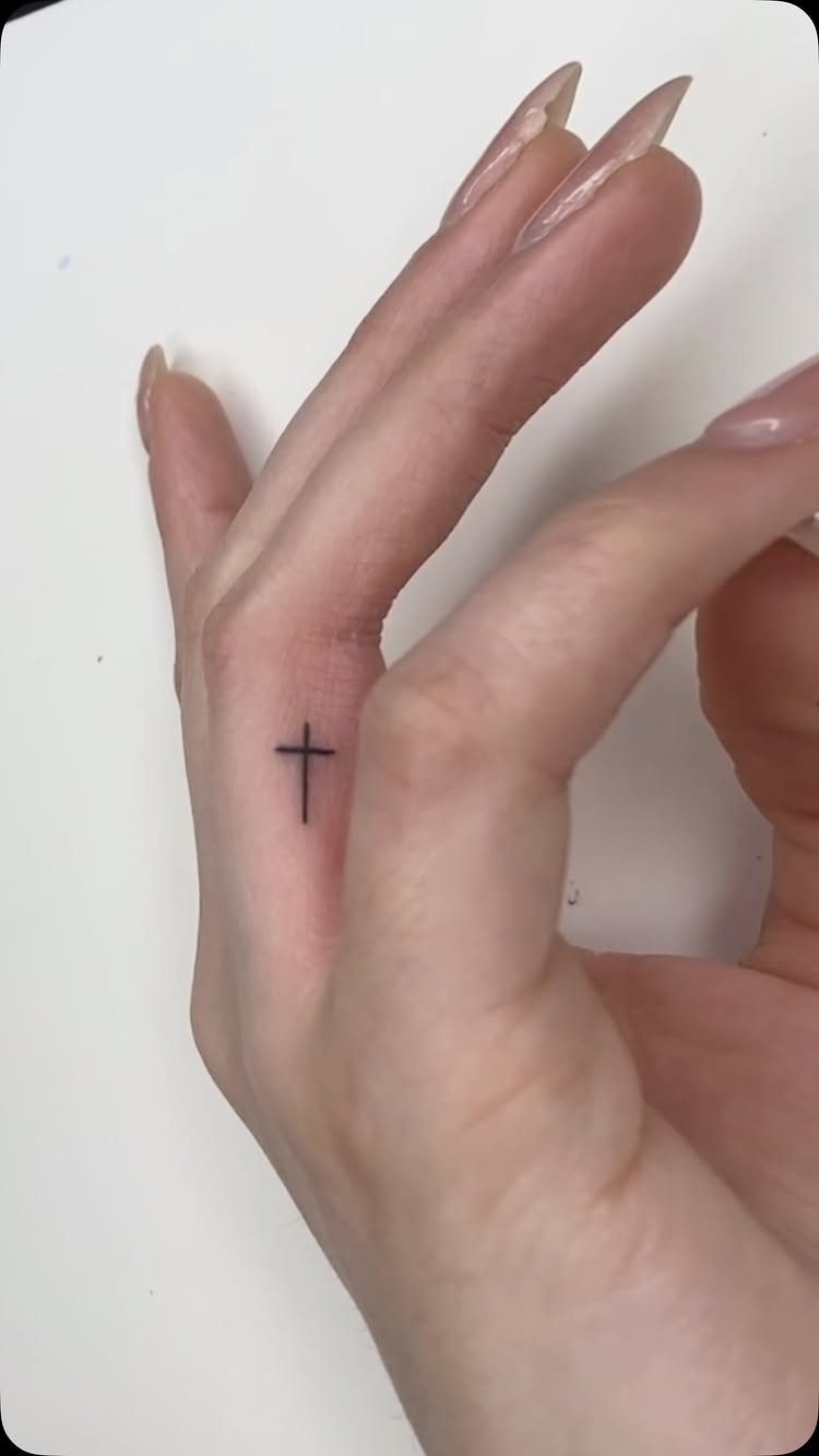 Small black cross tattoo on the side of a woman's ring finger