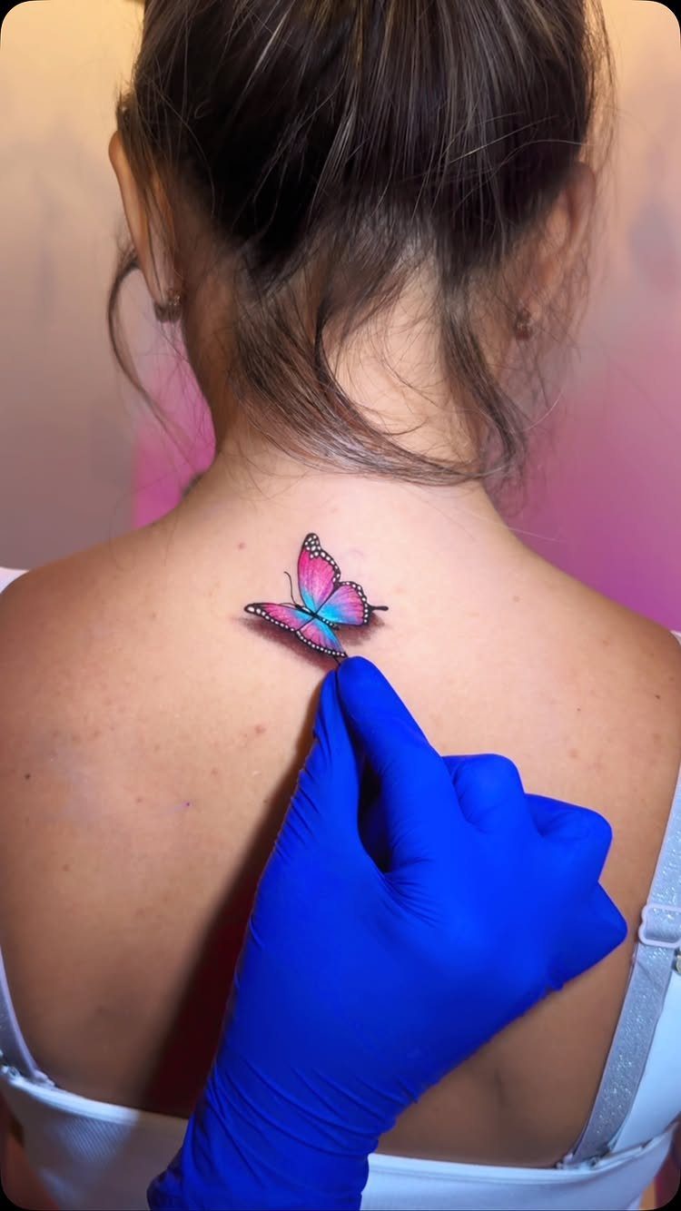 Colorful butterfly tattoo on a woman's upper back with a blue gloved hand pointing.