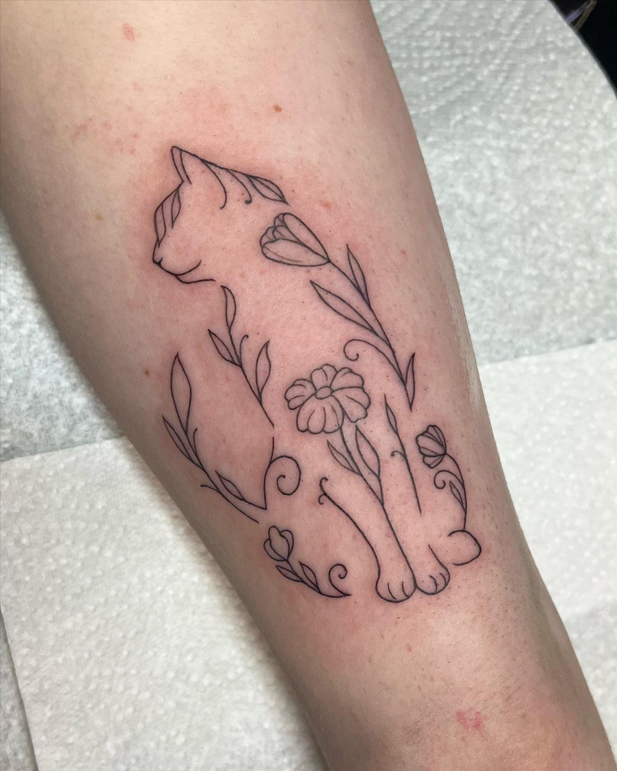 Fine-line tattoo of a sitting cat with flowers on a person's arm.