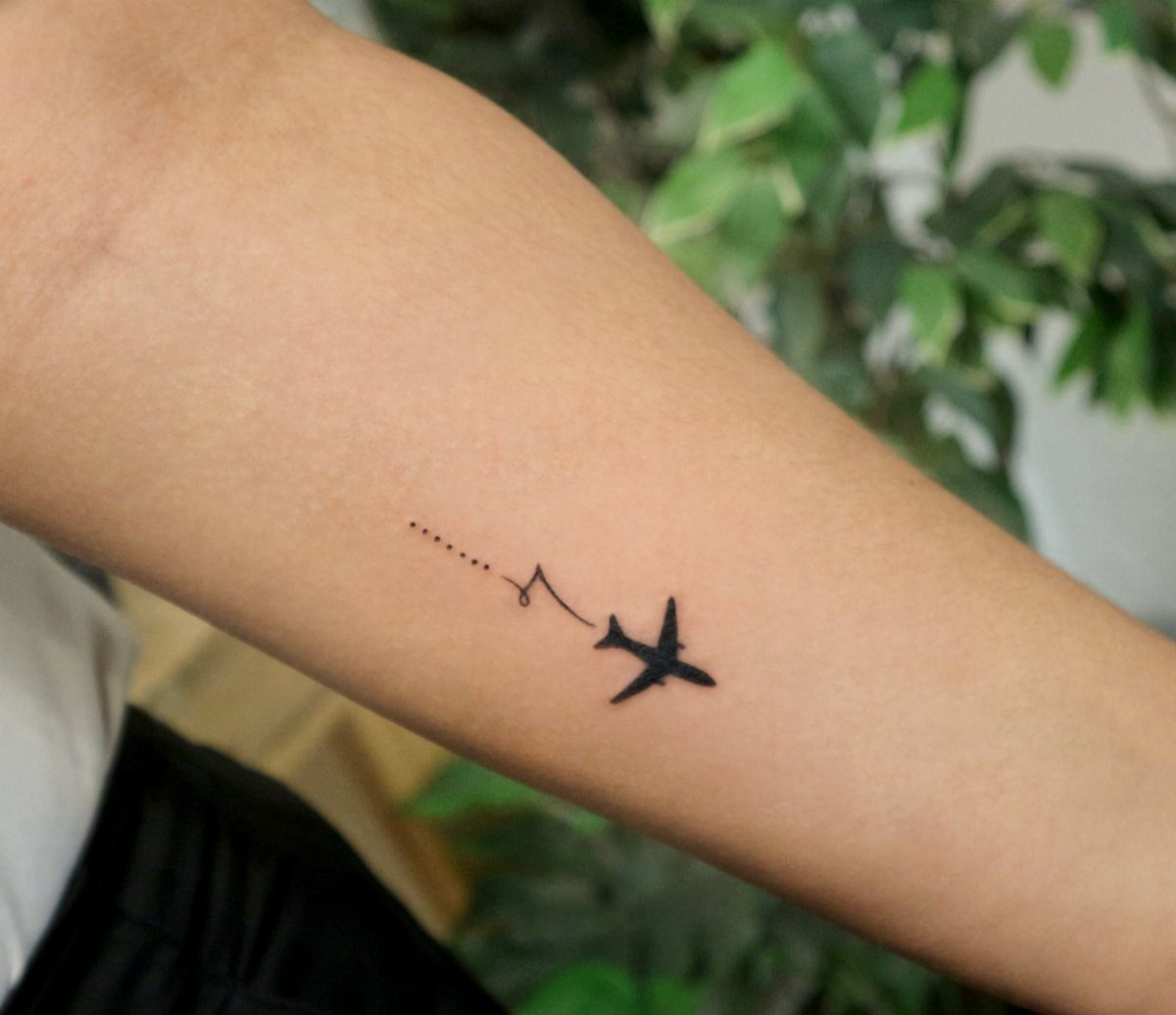 Small black airplane tattoo with a dotted trail on a person's forearm