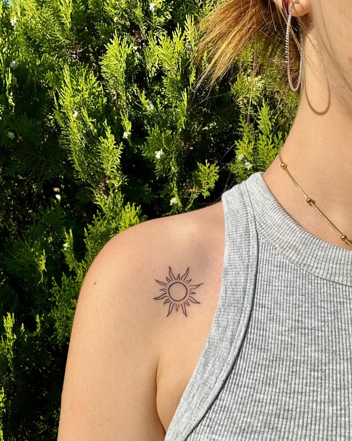 Small sun tattoo with rays on a person's shoulder against green foliage.
