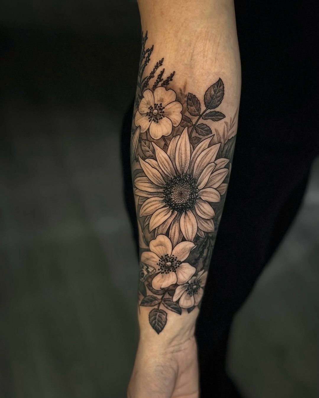 Floral forearm tattoo featuring a large sunflower and smaller blossoms in black and grey ink.
