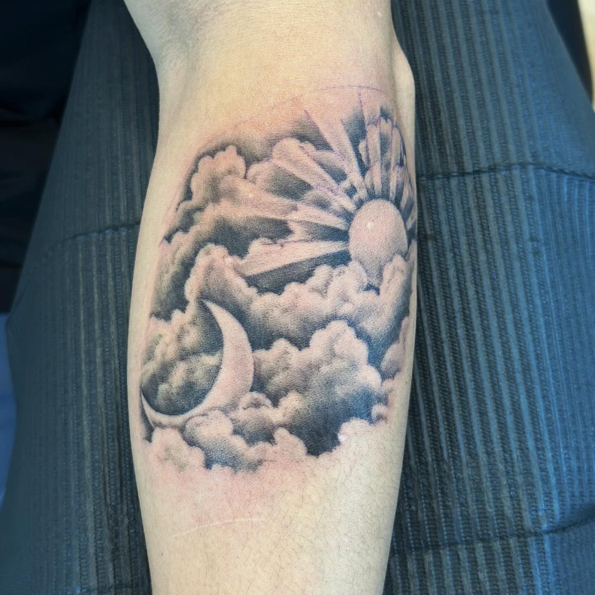 Sun and moon tattoo with clouds on a person's forearm in black and grey.