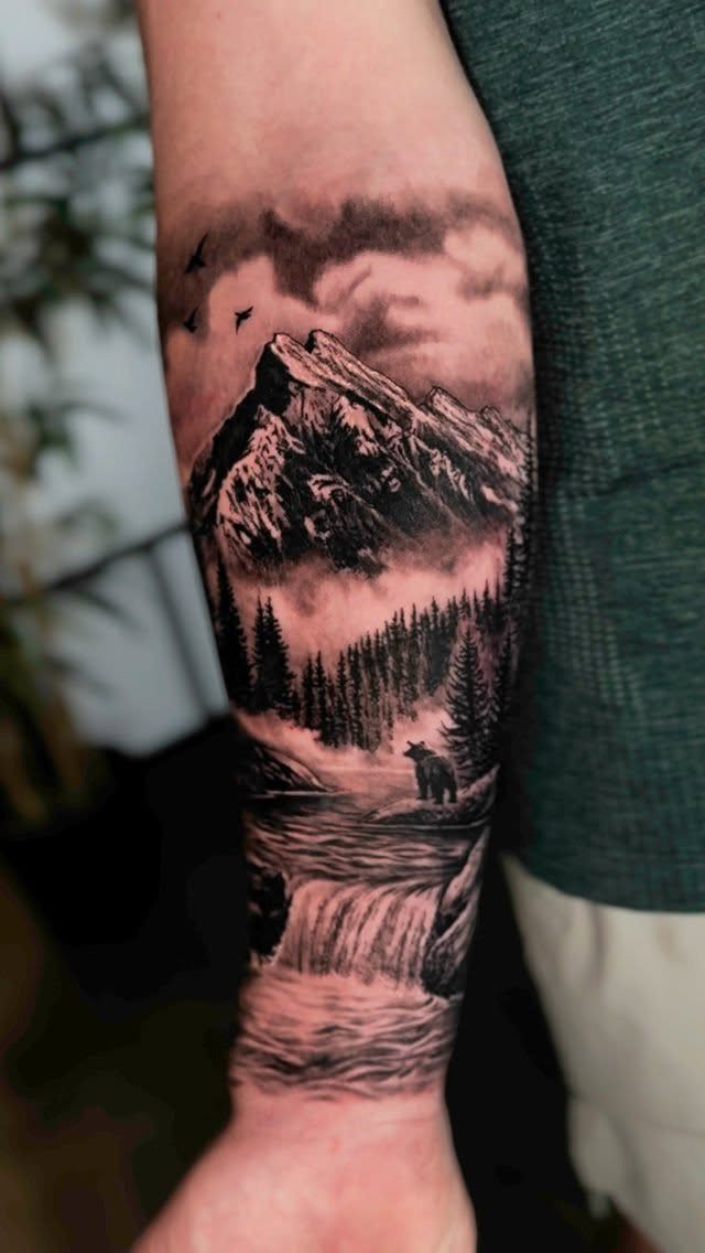 Forearm tattoo depicting a mountain landscape with a forest, river, and a bear.