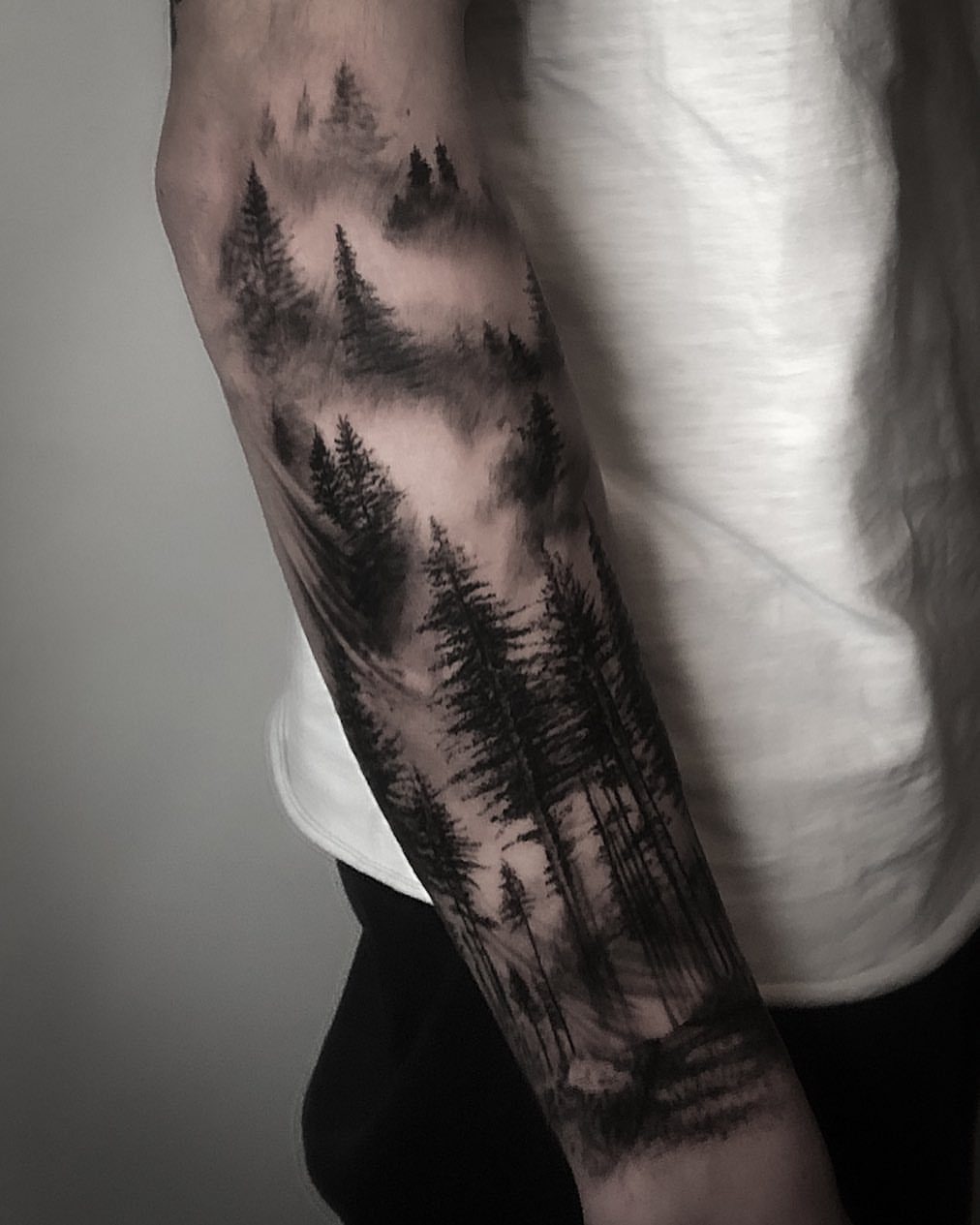Forearm tattoo featuring a detailed black and grey pine forest with misty atmosphere.