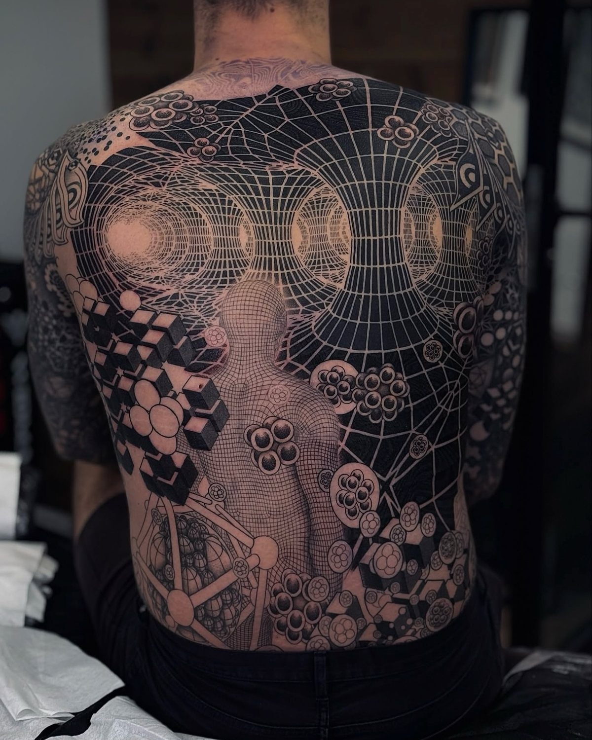 Man's back with a detailed geometric tattoo featuring grids, spheres, and abstract shapes.
