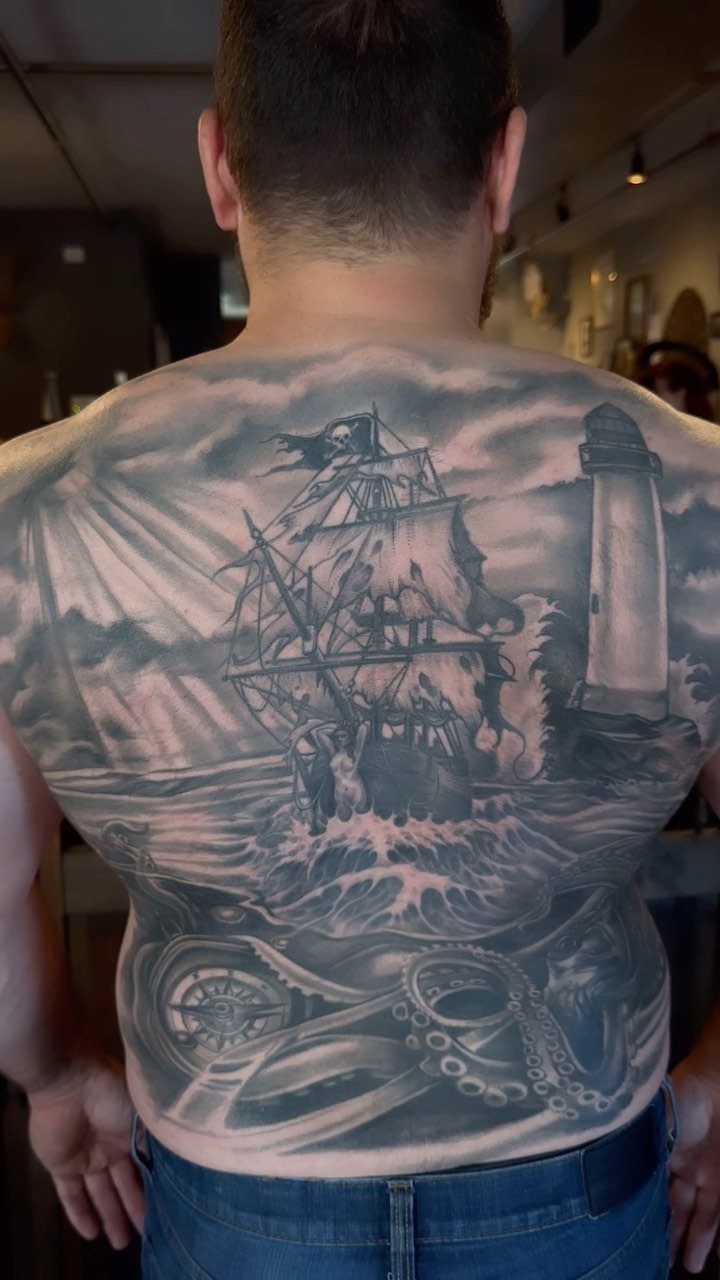 Full back tattoo of a sailing ship, lighthouse, and nautical compass in grey ink.