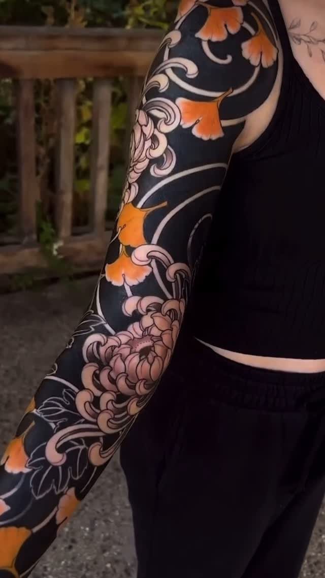 Full sleeve tattoo with pink chrysanthemums and orange ginkgo leaves on an arm.