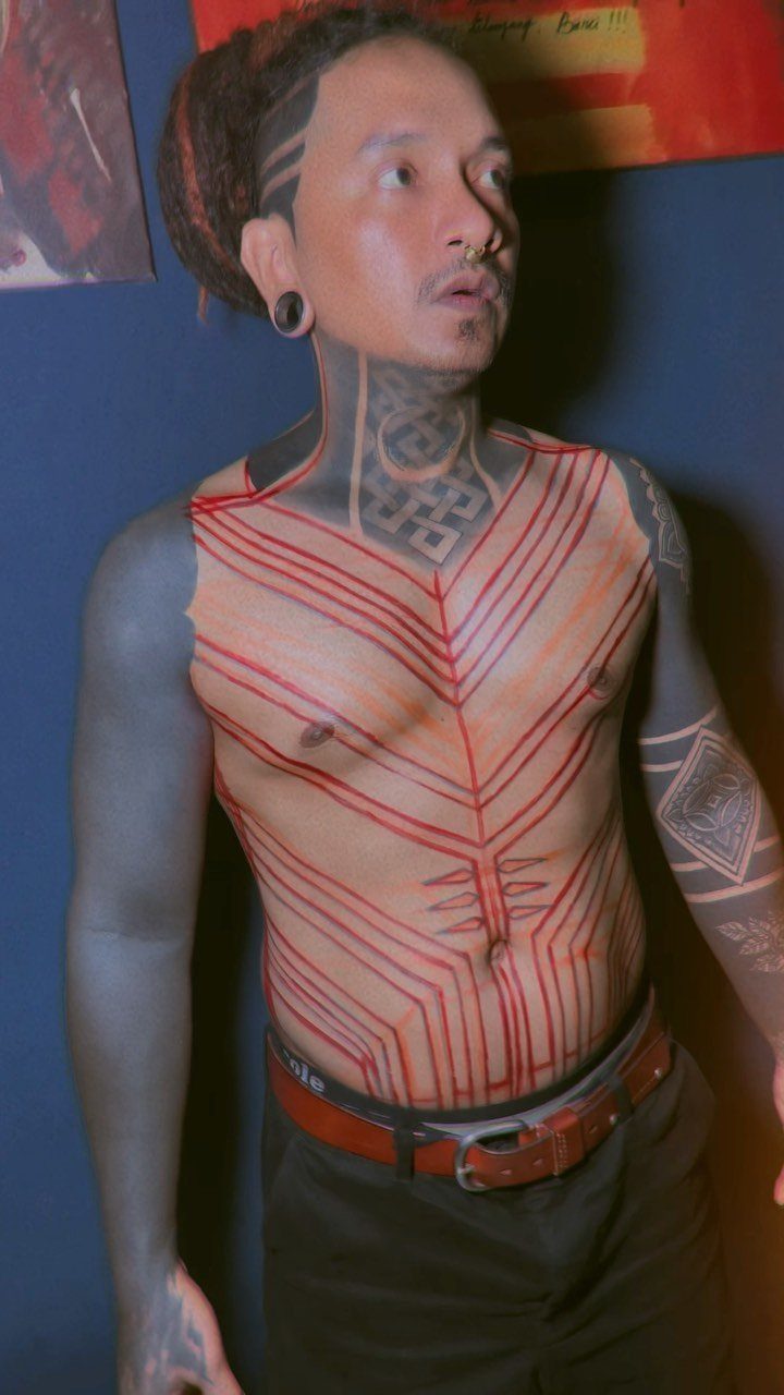 Man with red and black geometric tattoos covering his chest, neck, and arms.