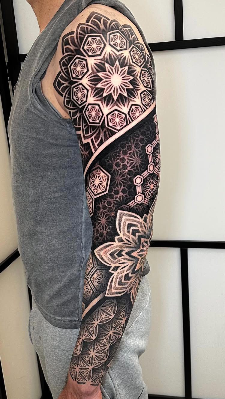 Man's arm with a full sleeve tattoo featuring intricate black geometric mandala patterns.