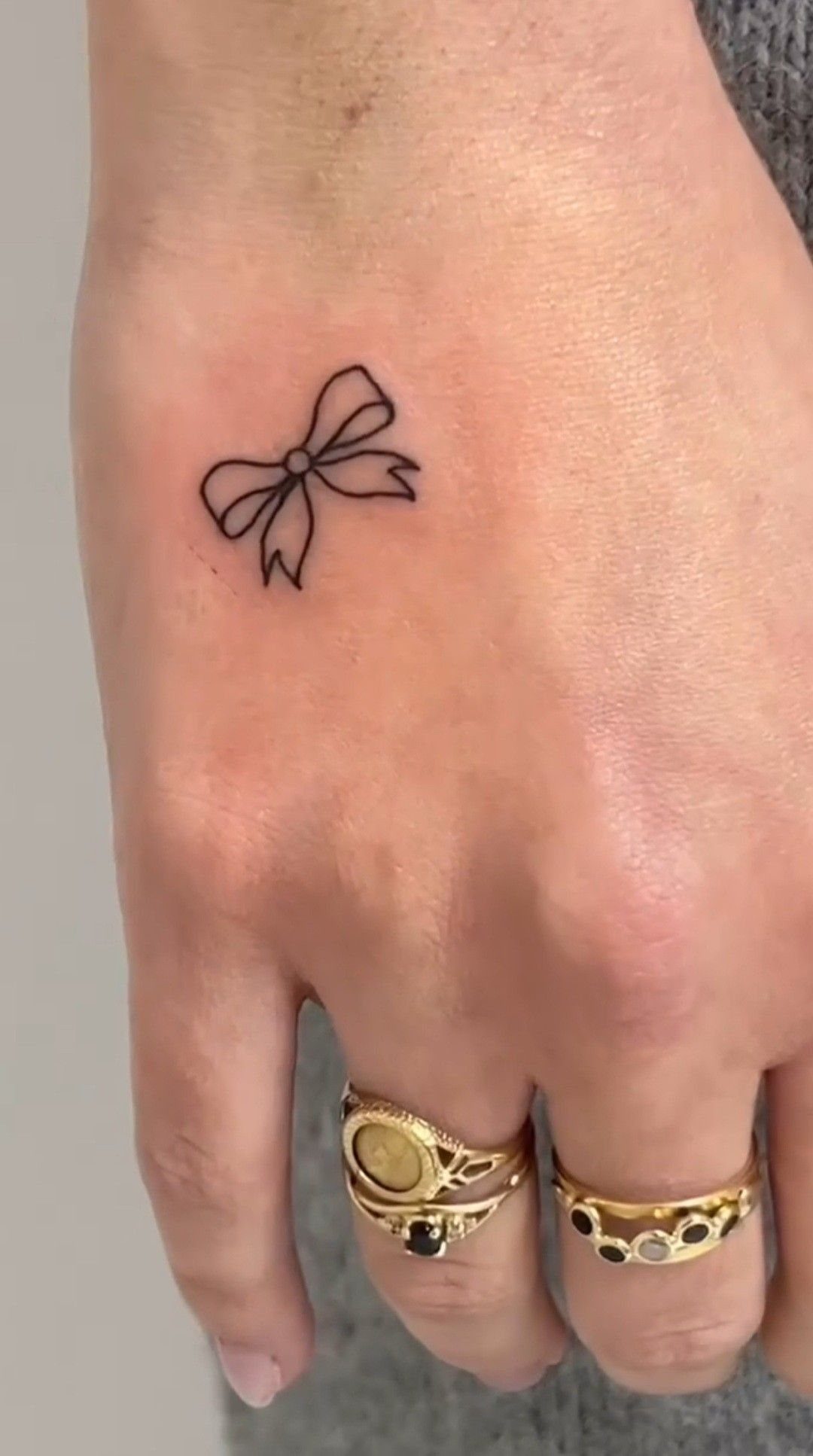 Small black ink bow tattoo on the back of a hand with gold rings