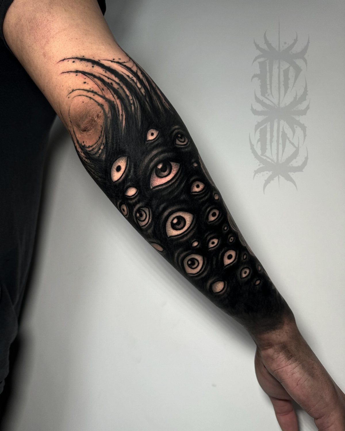 Forearm tattoo with heavy black ink featuring multiple surreal eyes and swirling patterns.