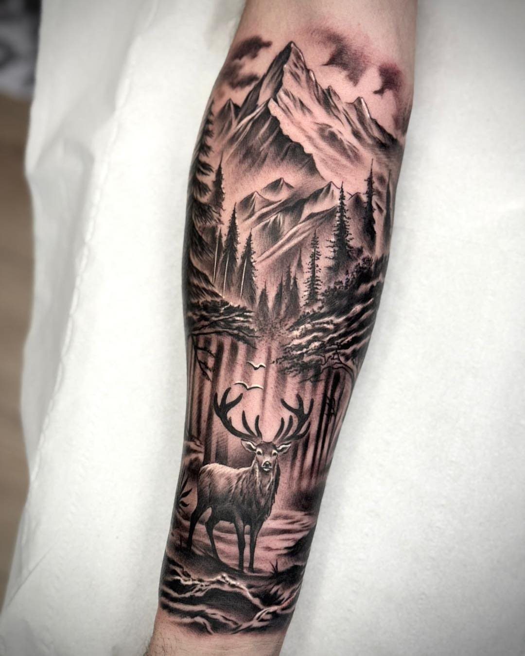Forearm half sleeve tattoo depicting a stag in a forest with mountains.