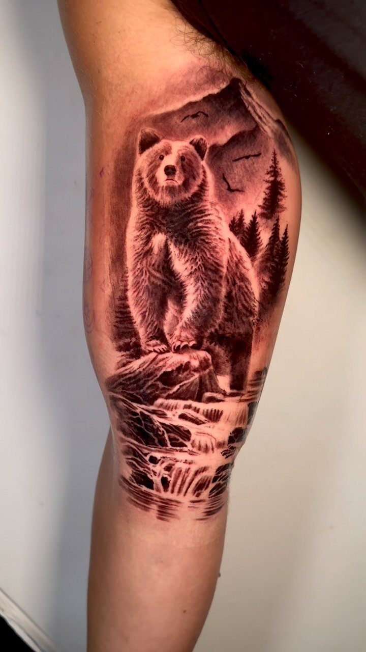 Bear tattoo on forearm with mountains, pine trees, and a flowing waterfall.