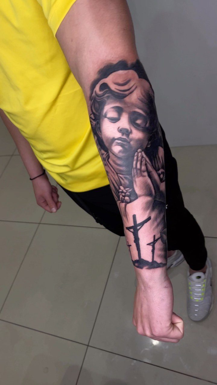 Forearm tattoo of a praying cherub and three crosses in black and grey ink.