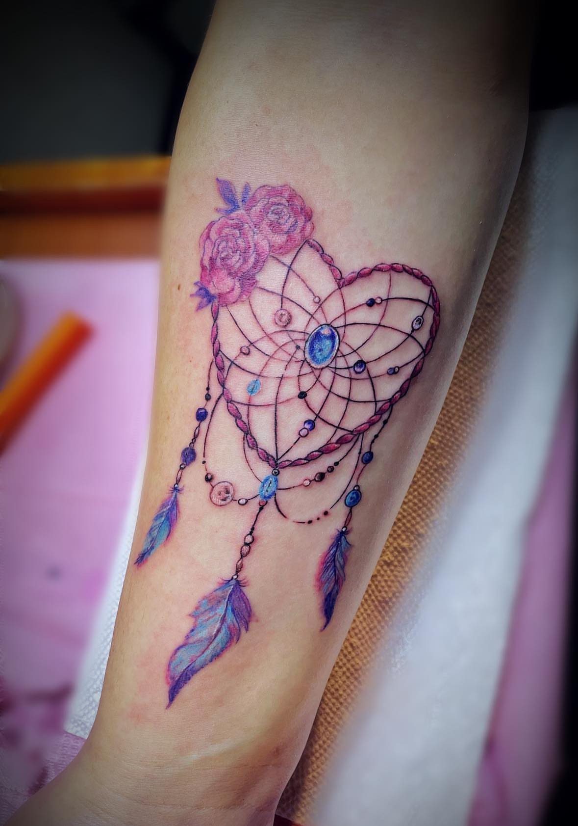 Heart-shaped dreamcatcher tattoo with purple roses and blue feathers on a forearm.