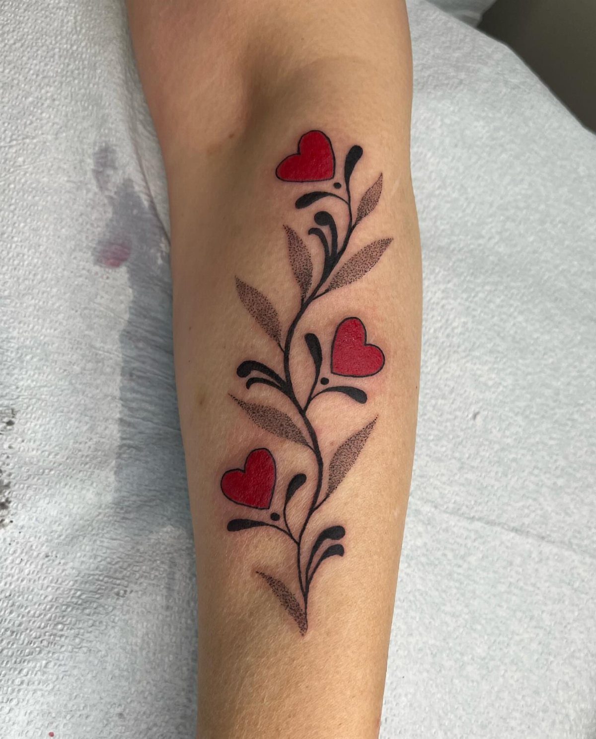 Tattoo on a forearm featuring a black vine with leaves and three red hearts.