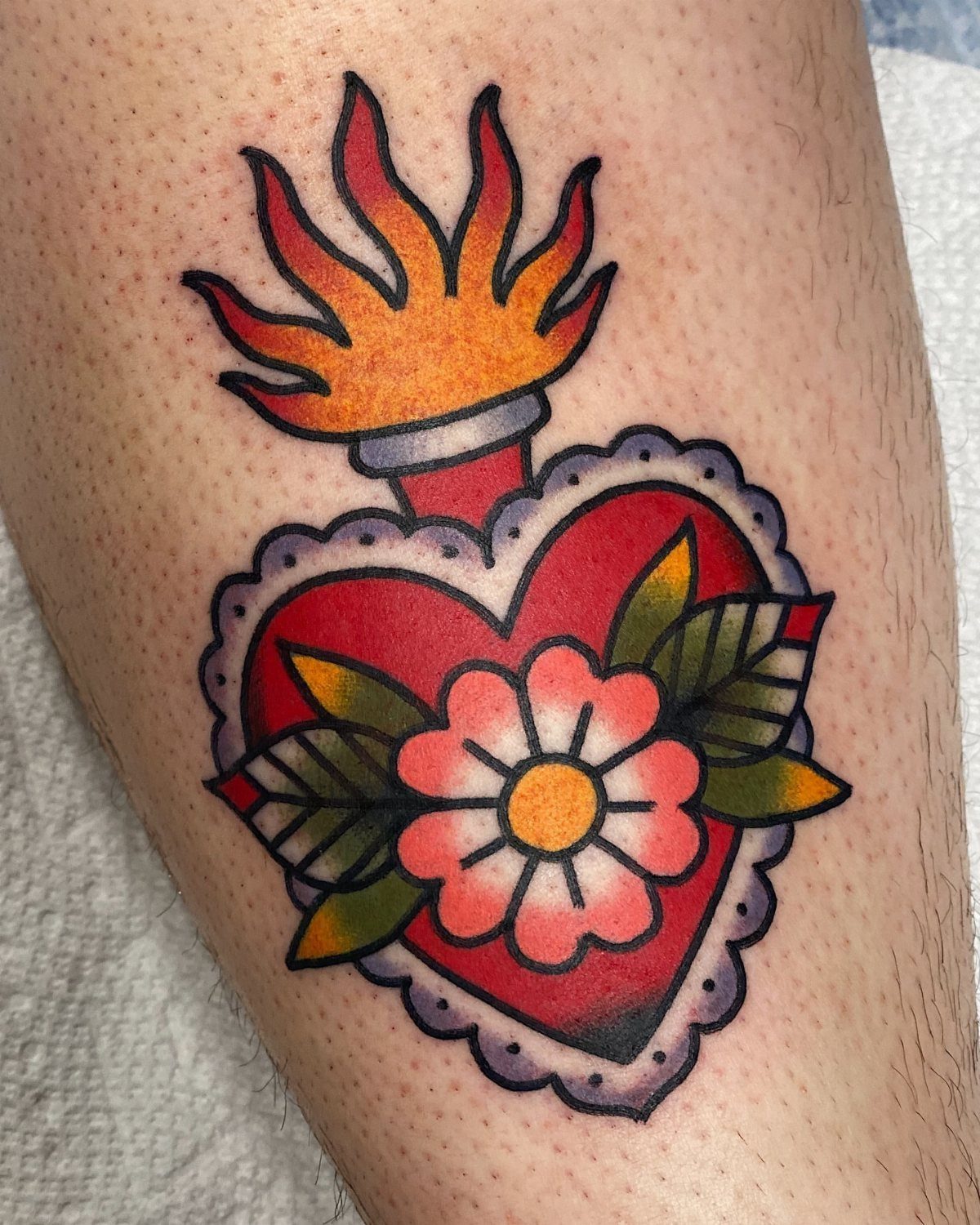 Traditional red heart tattoo with a pink flower and orange flames on skin.