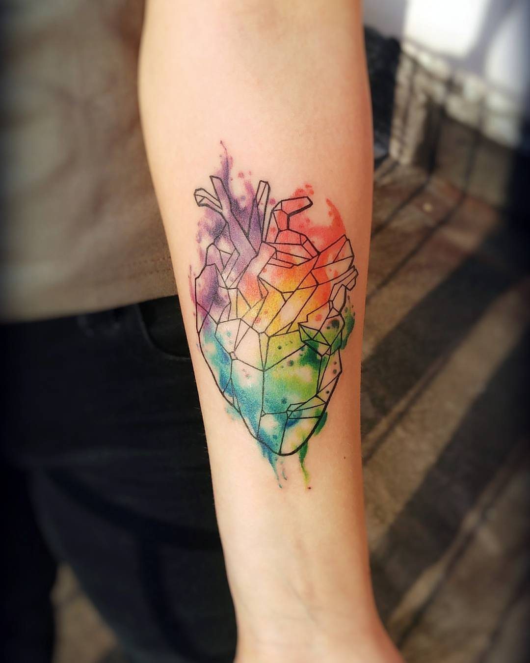 Anatomical heart tattoo with geometric lines and vibrant watercolor fills on a forearm.