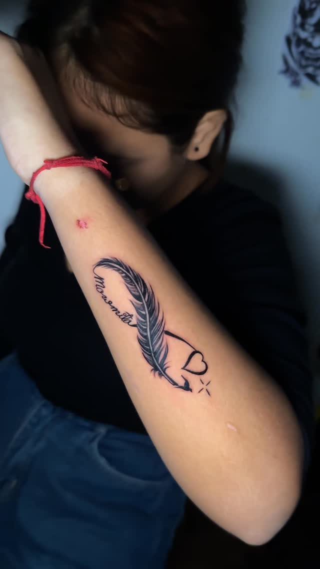 Feather tattoo with a small heart and cursive text on a woman's forearm.