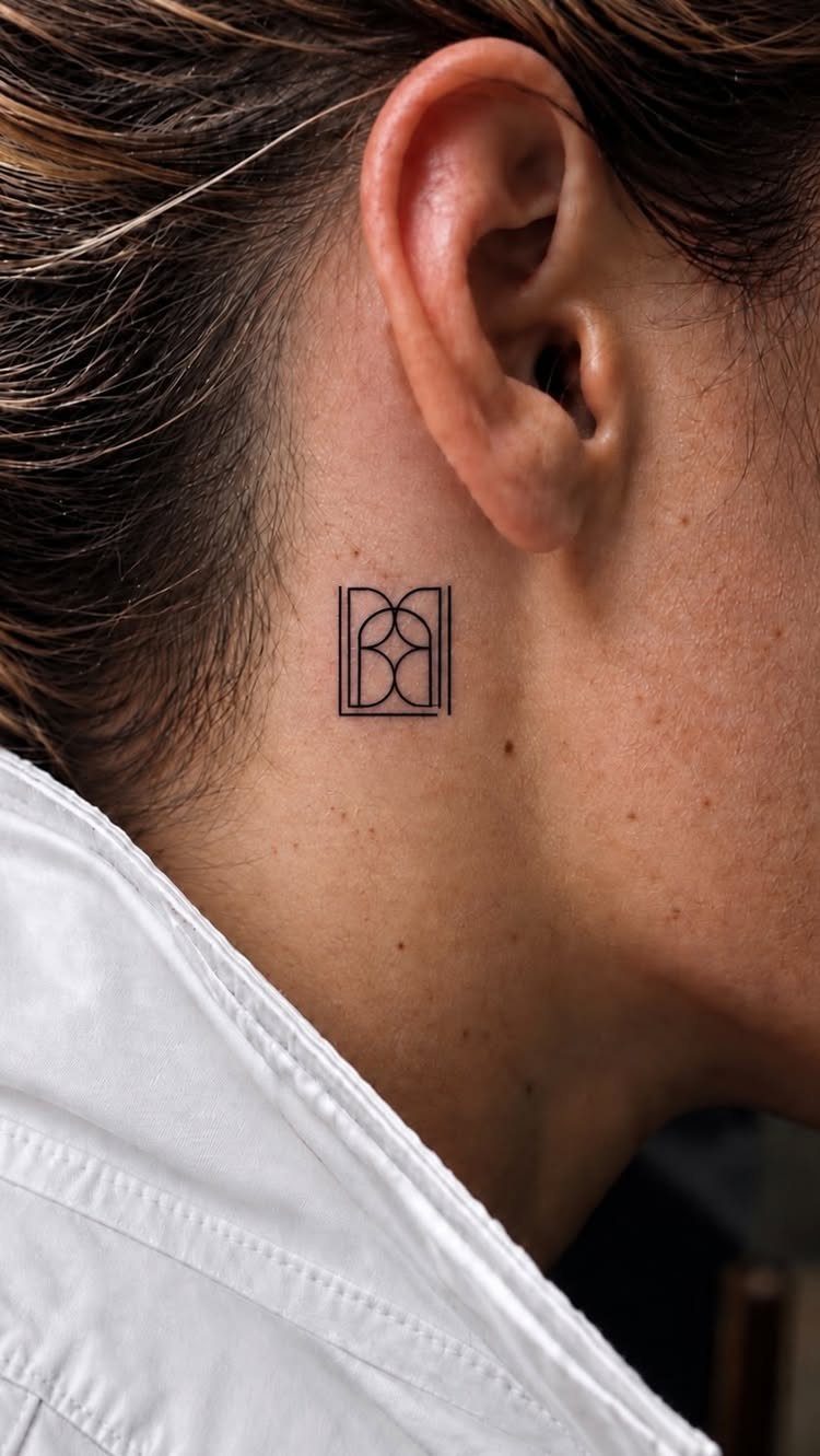 Geometric monogram tattoo on the side of a person's neck behind the ear