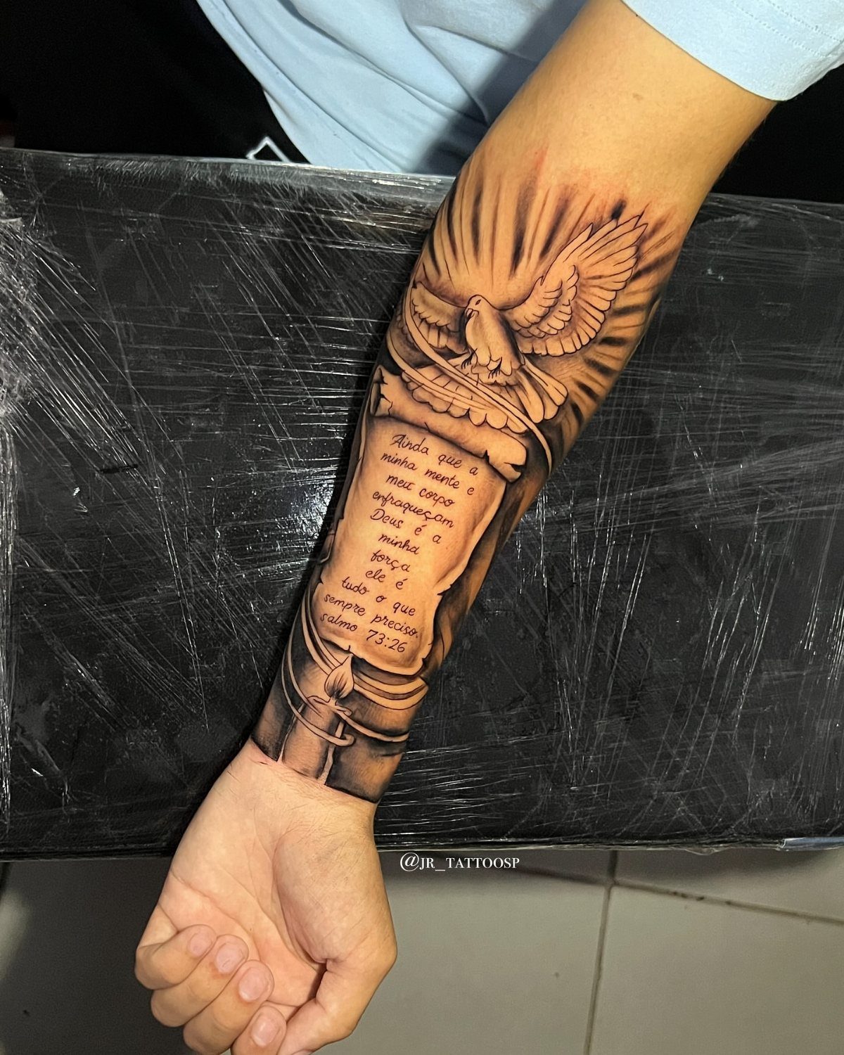 Inner arm tattoo of a dove above a scroll with religious scripture text.
