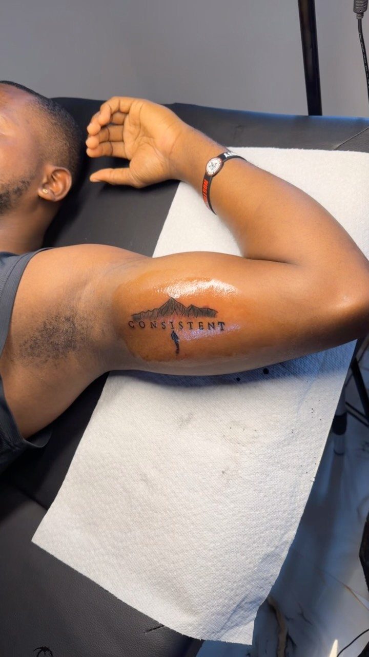 Man's inner bicep tattoo featuring the word CONSISTENT with a mountain peak silhouette.
