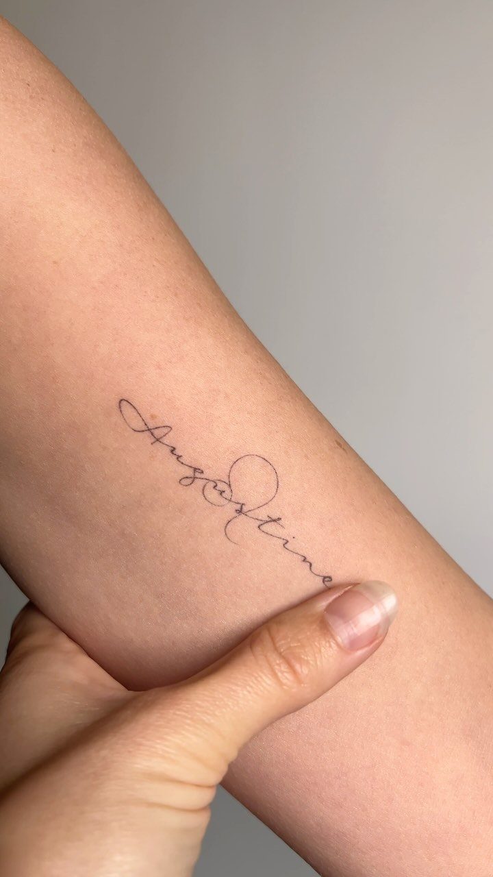 Cursive script tattoo reading Augustine on a person's inner forearm skin.