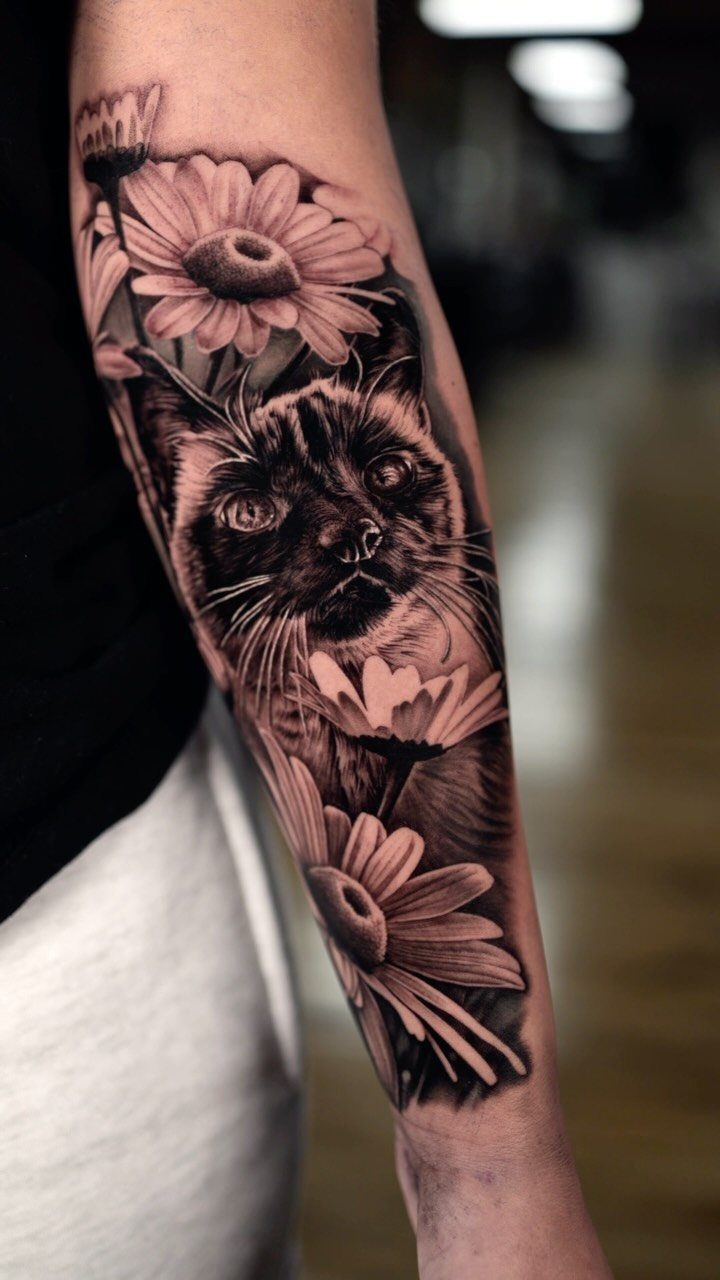 Inner forearm tattoo of a cat's face surrounded by blooming daisy flowers.