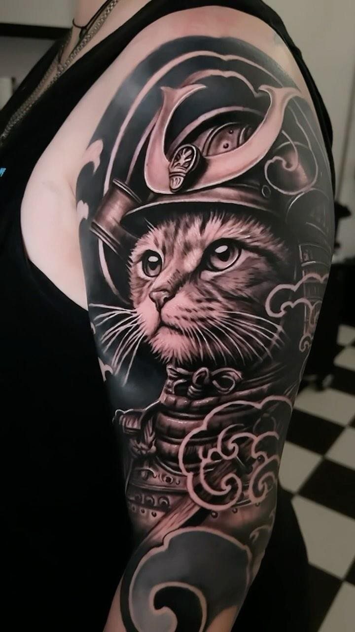Cat wearing samurai armor tattoo on a person's upper arm in black and grey.