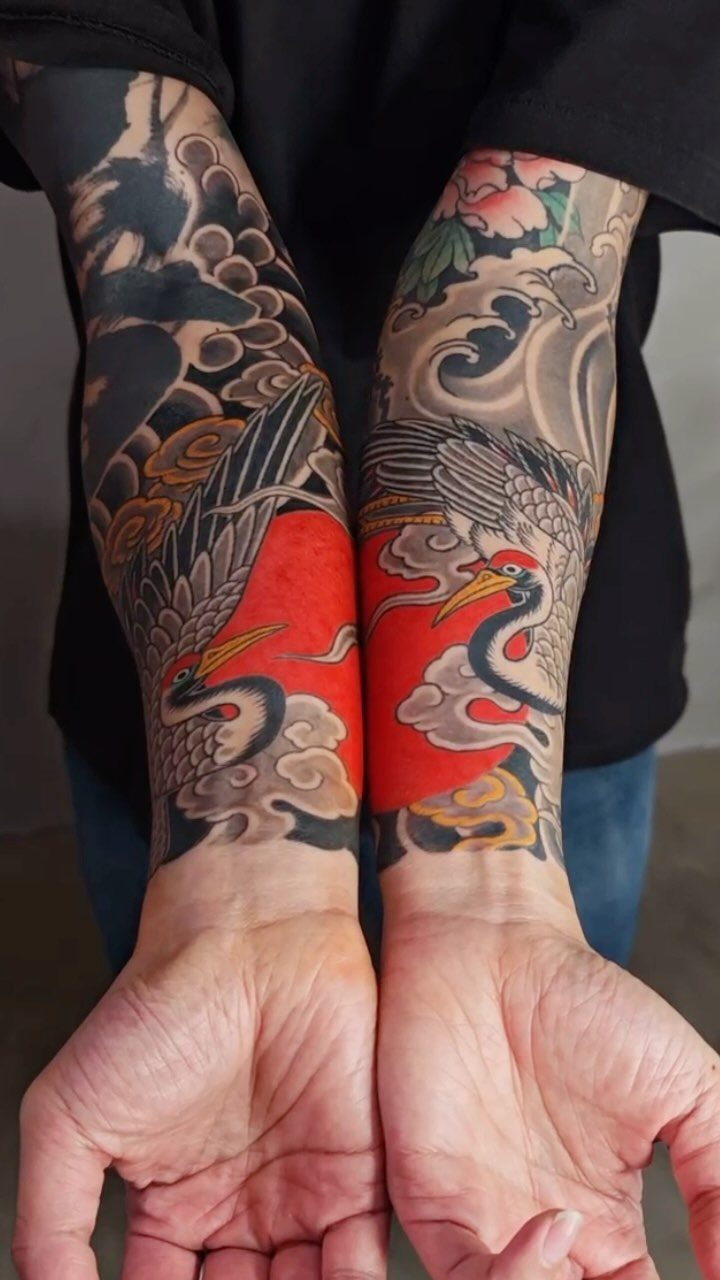 Japanese crane tattoos on two forearms with red backgrounds and grey waves.
