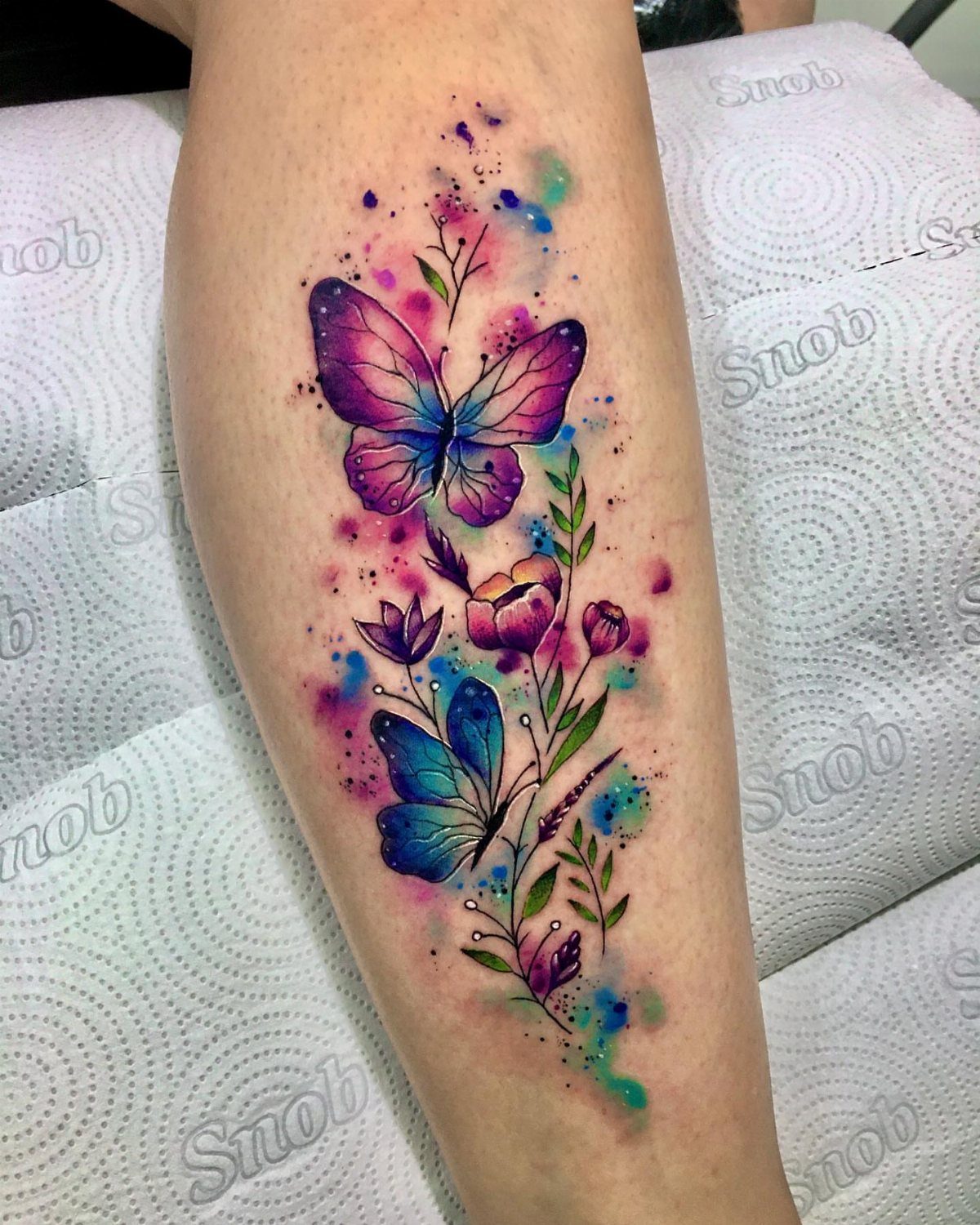 Watercolor tattoo of purple and blue butterflies with flowers on a person's leg