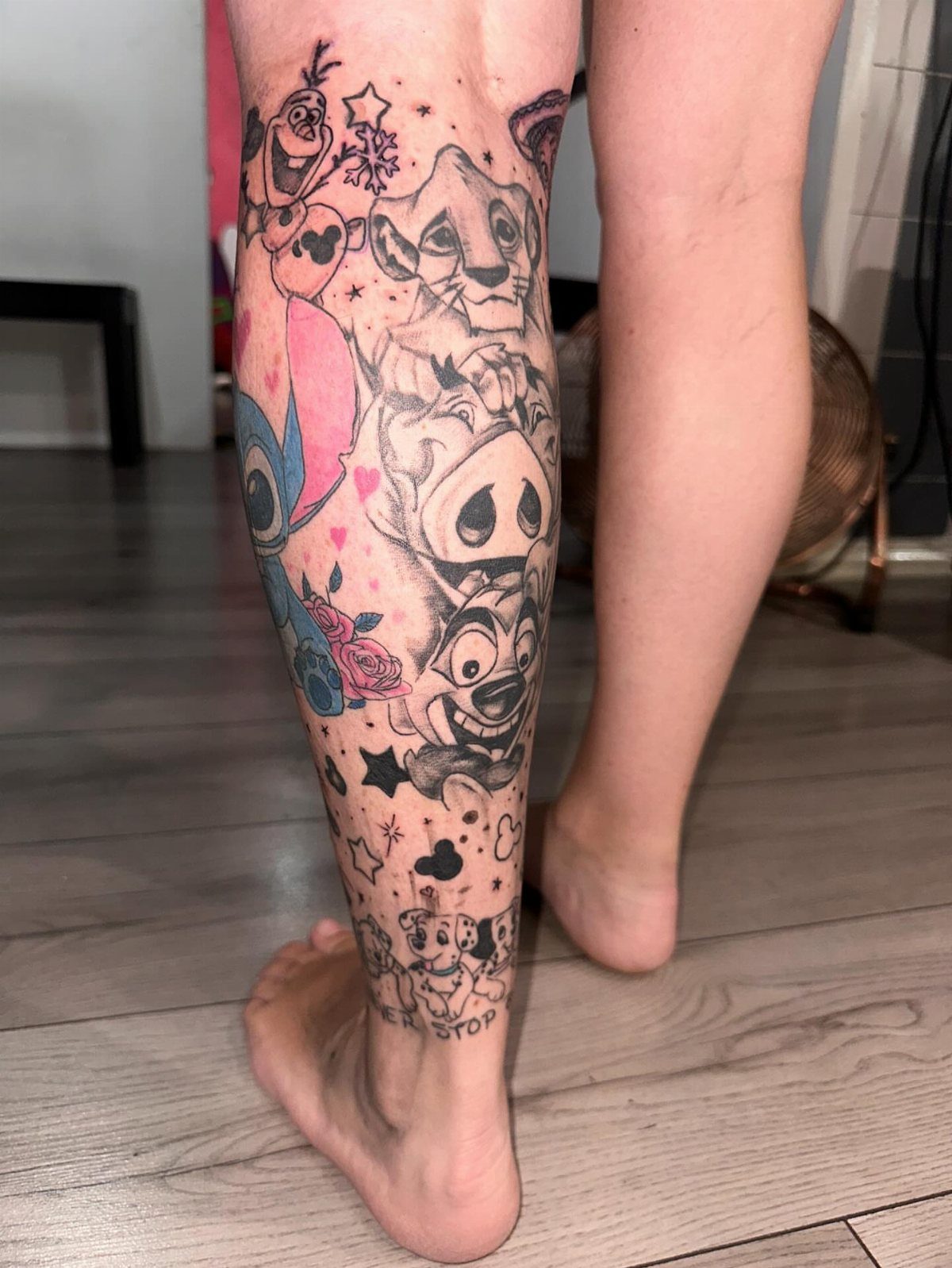 Lower leg with a colorful tattoo of various Disney characters on a wooden floor.