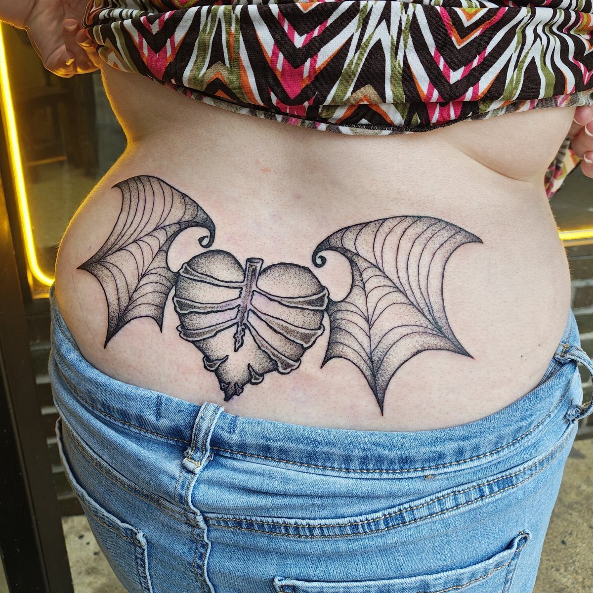 Heart with bat wings tattoo on a person's lower back above blue jeans