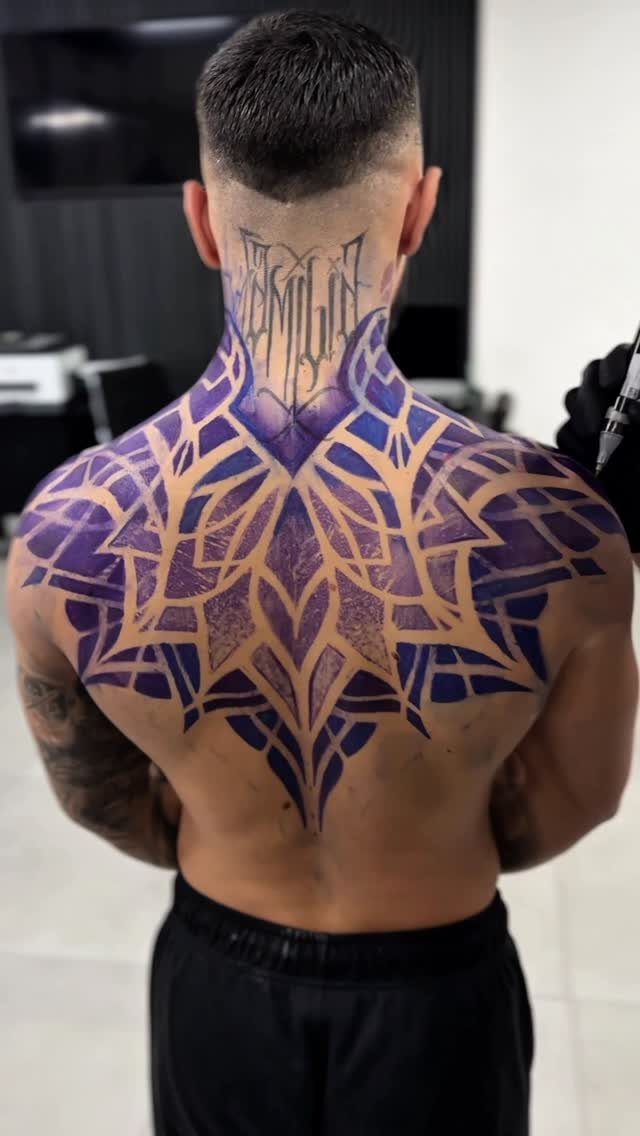 Geometric mandala tattoo in purple and gold covering a man's upper back and shoulders.