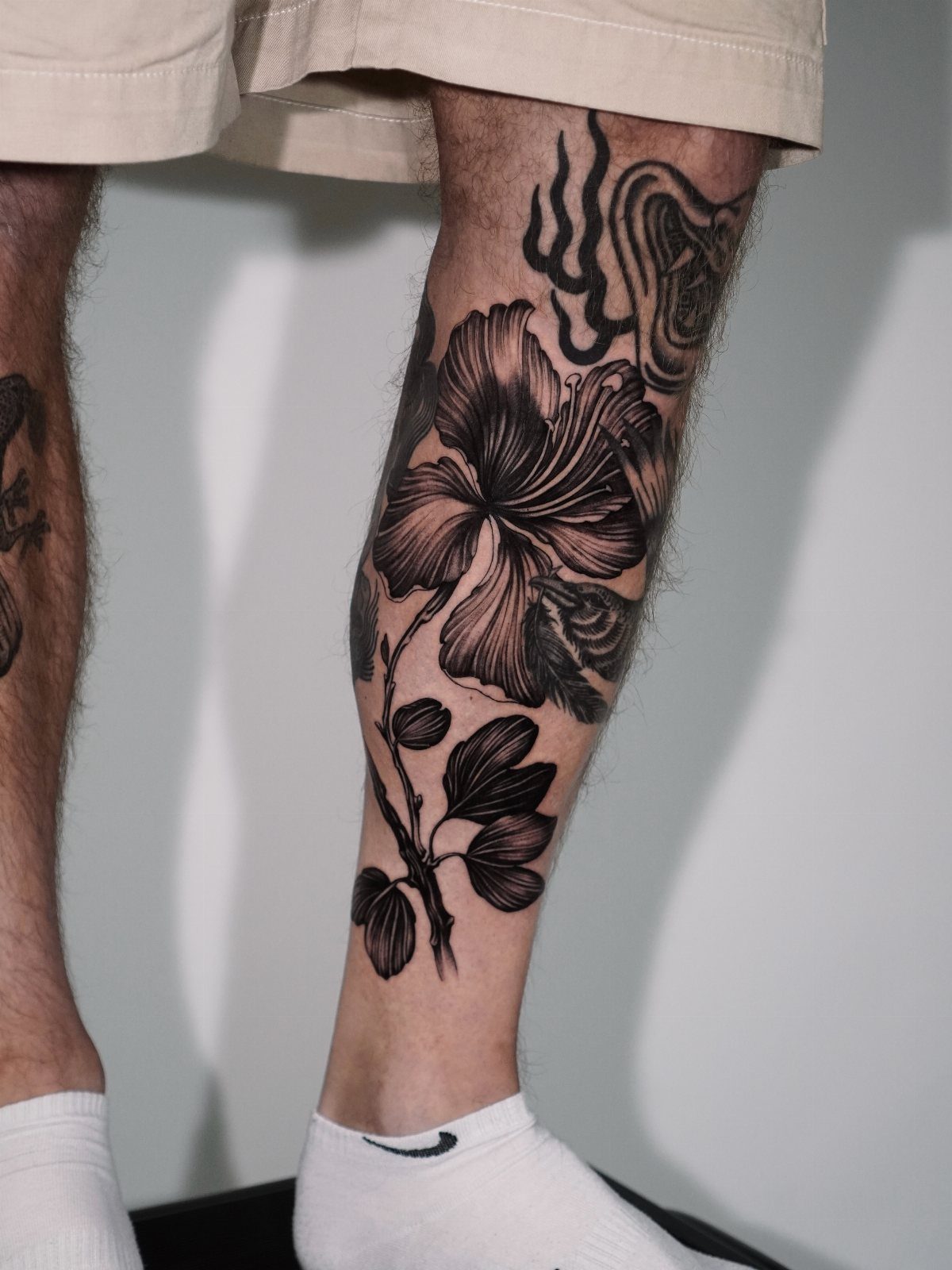 Man's calf with a detailed black ink botanical hibiscus flower tattoo.