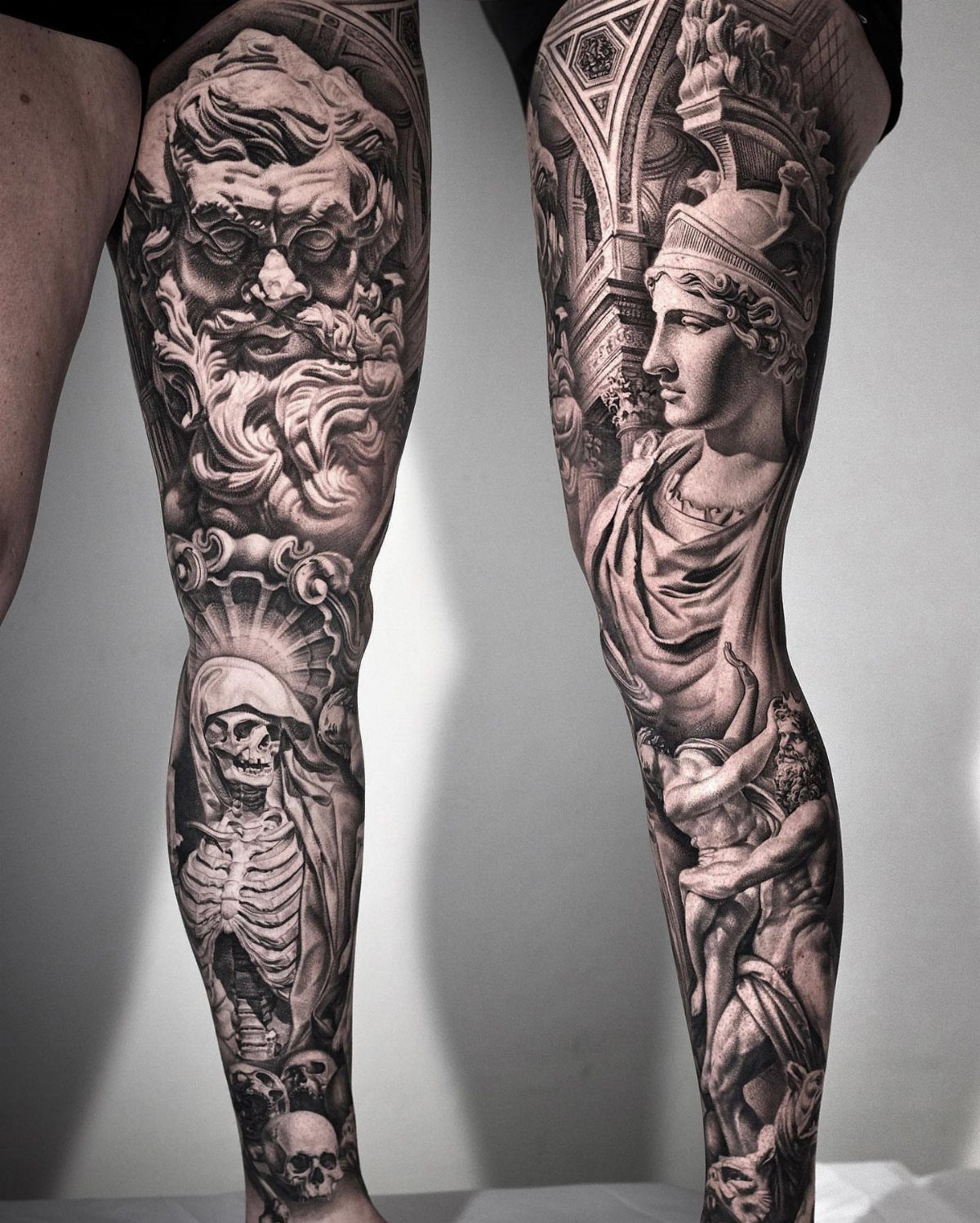 Detailed black and grey leg tattoos featuring classical figures and a skeleton.