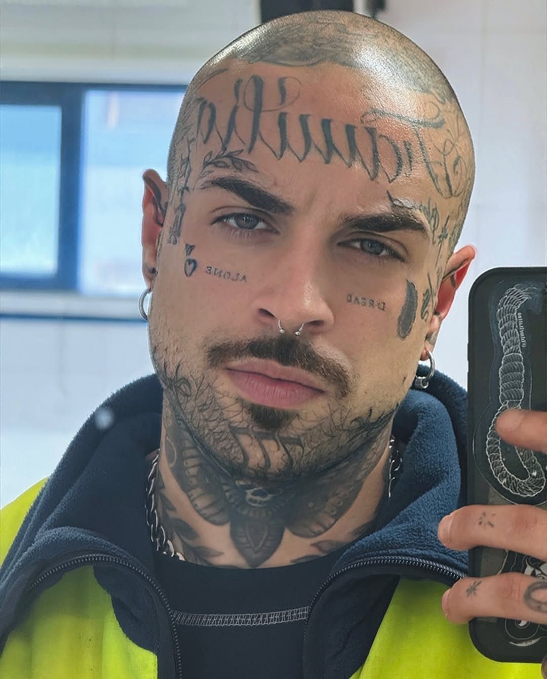 Bald man with extensive black ink tattoos on his face and neck.