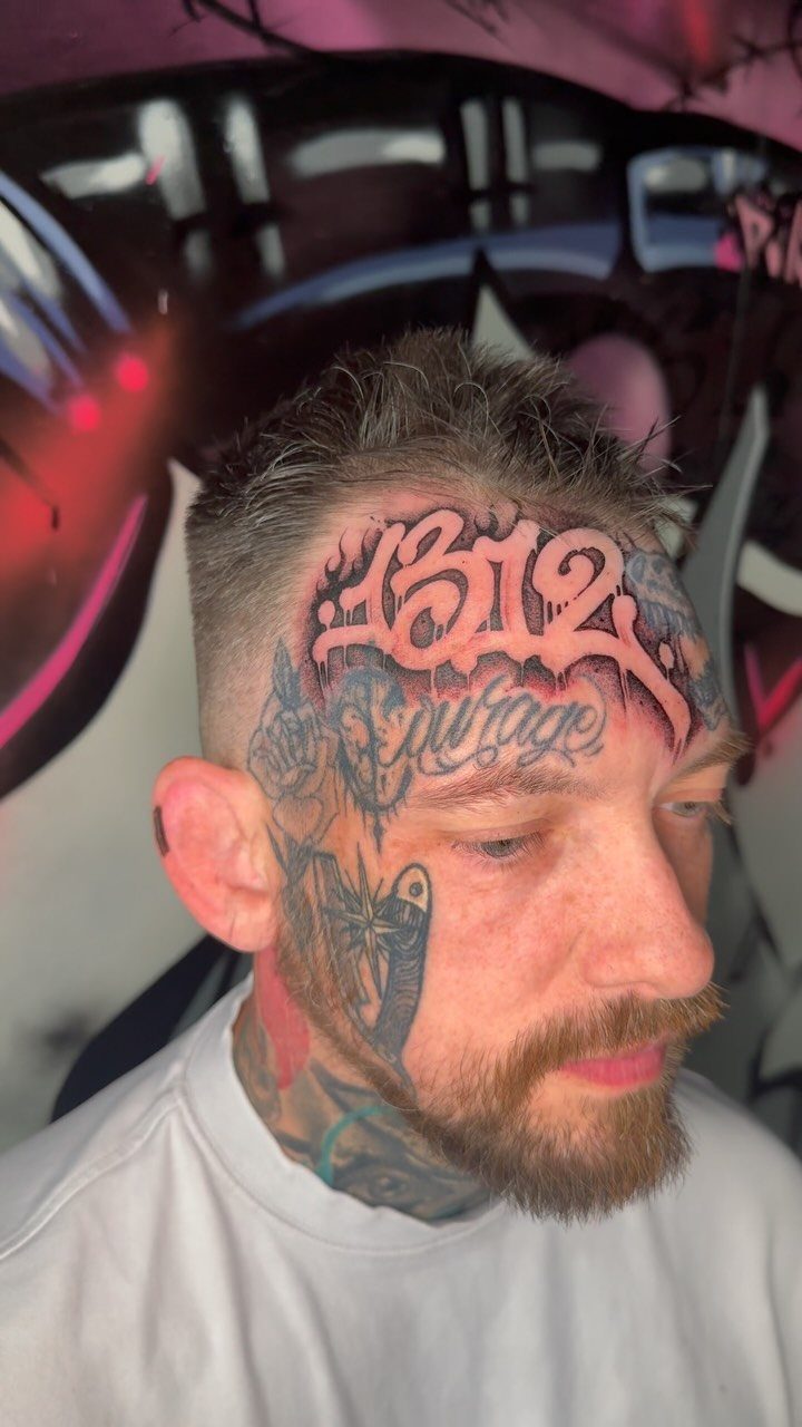 Man with graffiti script tattoos on his forehead and neck against a graffiti wall.