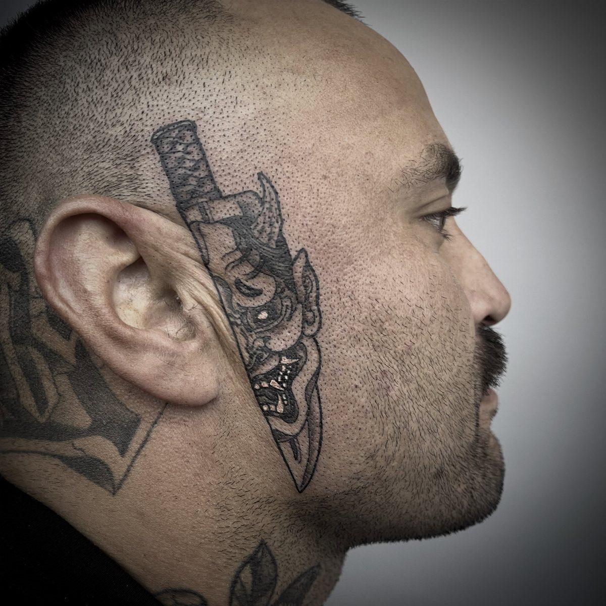Man's cheek tattoo featuring a dagger with an integrated Oni mask design.