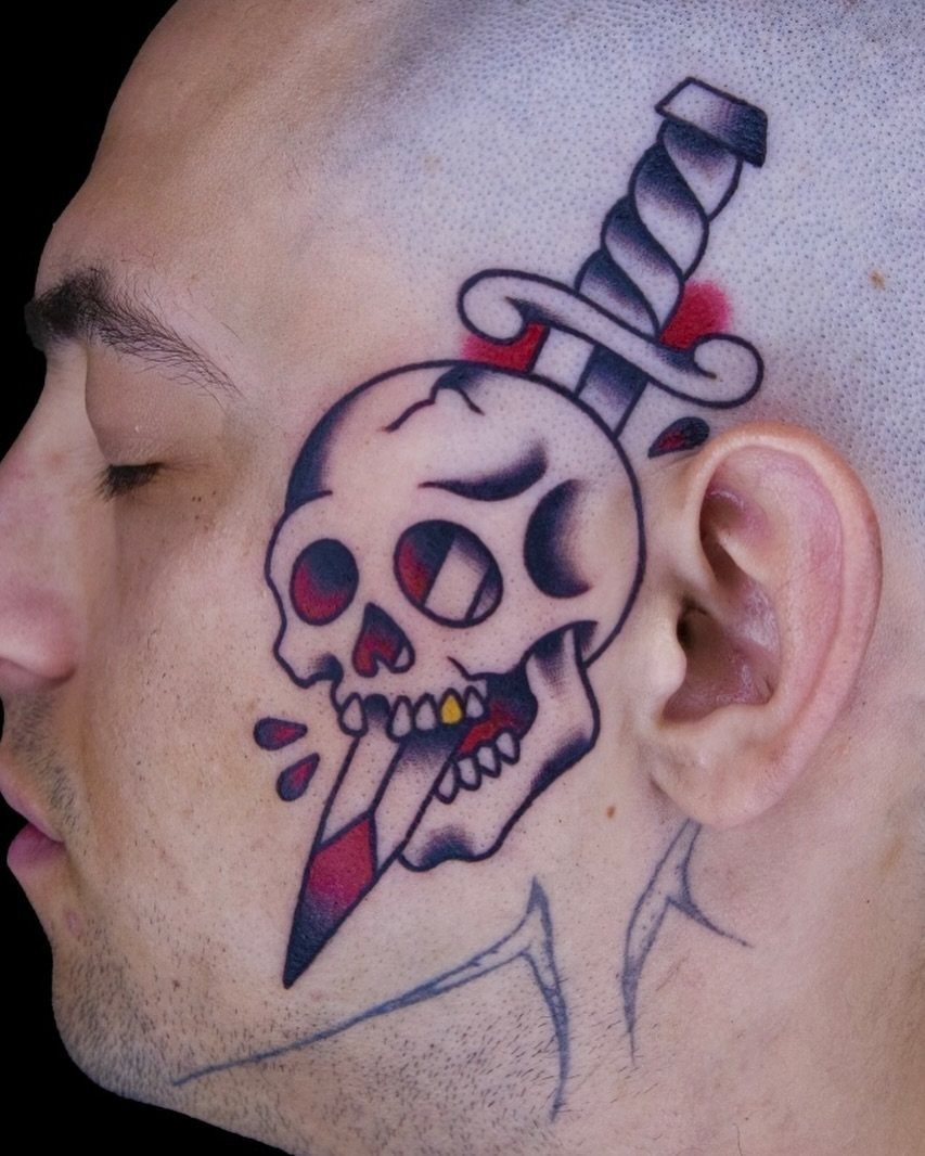 Man's face with a skull and dagger tattoo on his cheek and temple.
