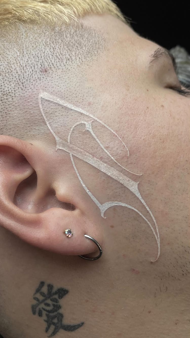 White ink stylized sigil tattoo on a man's cheek and temple area.