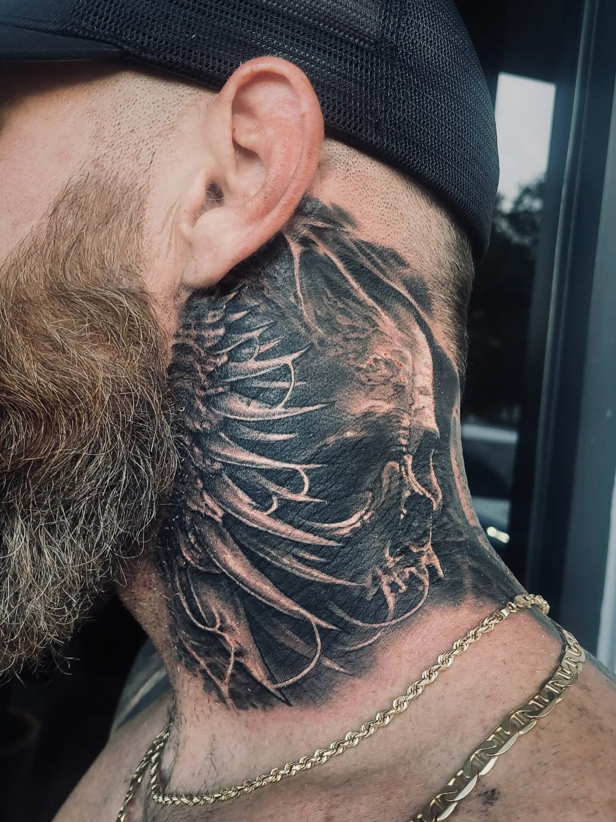 Man's neck with a detailed black and grey skull tattoo and reddish-brown beard.