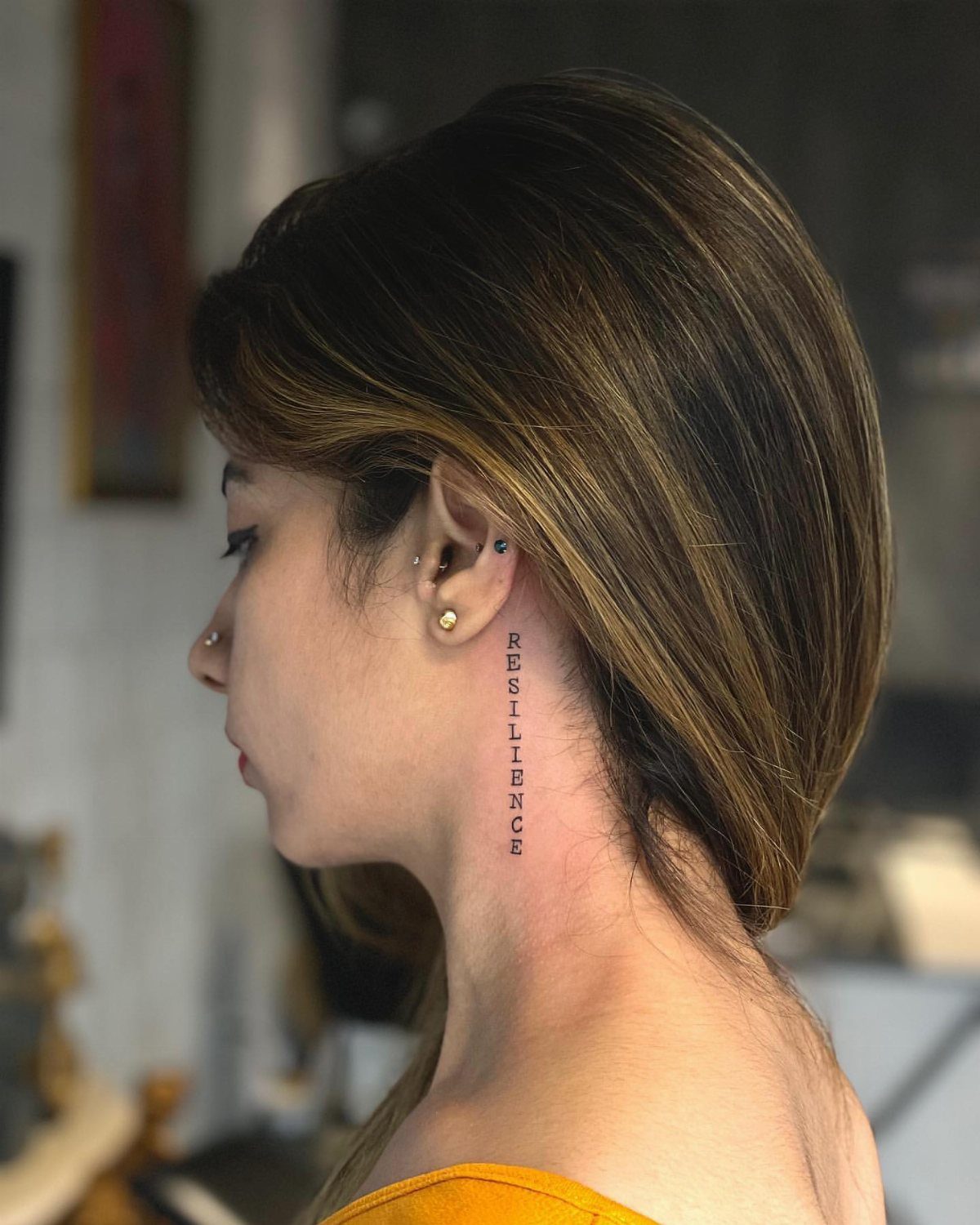 Woman with a vertical text tattoo reading RESILIENCE on the side of her neck.
